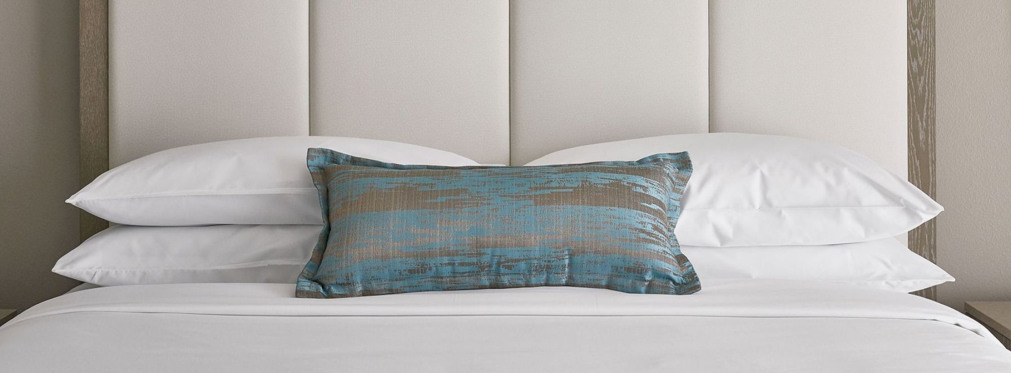 A neatly made bed with white pillows and a blue and brown accent pillow is shown against a light-colored headboard.