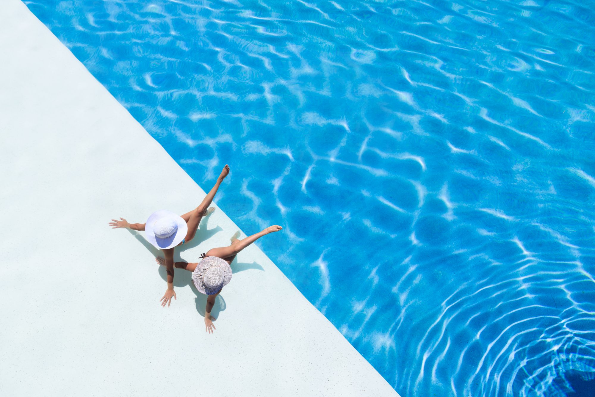 Two people wearing hats sit by the edge of a bright blue swimming pool, with legs dangling in the water.