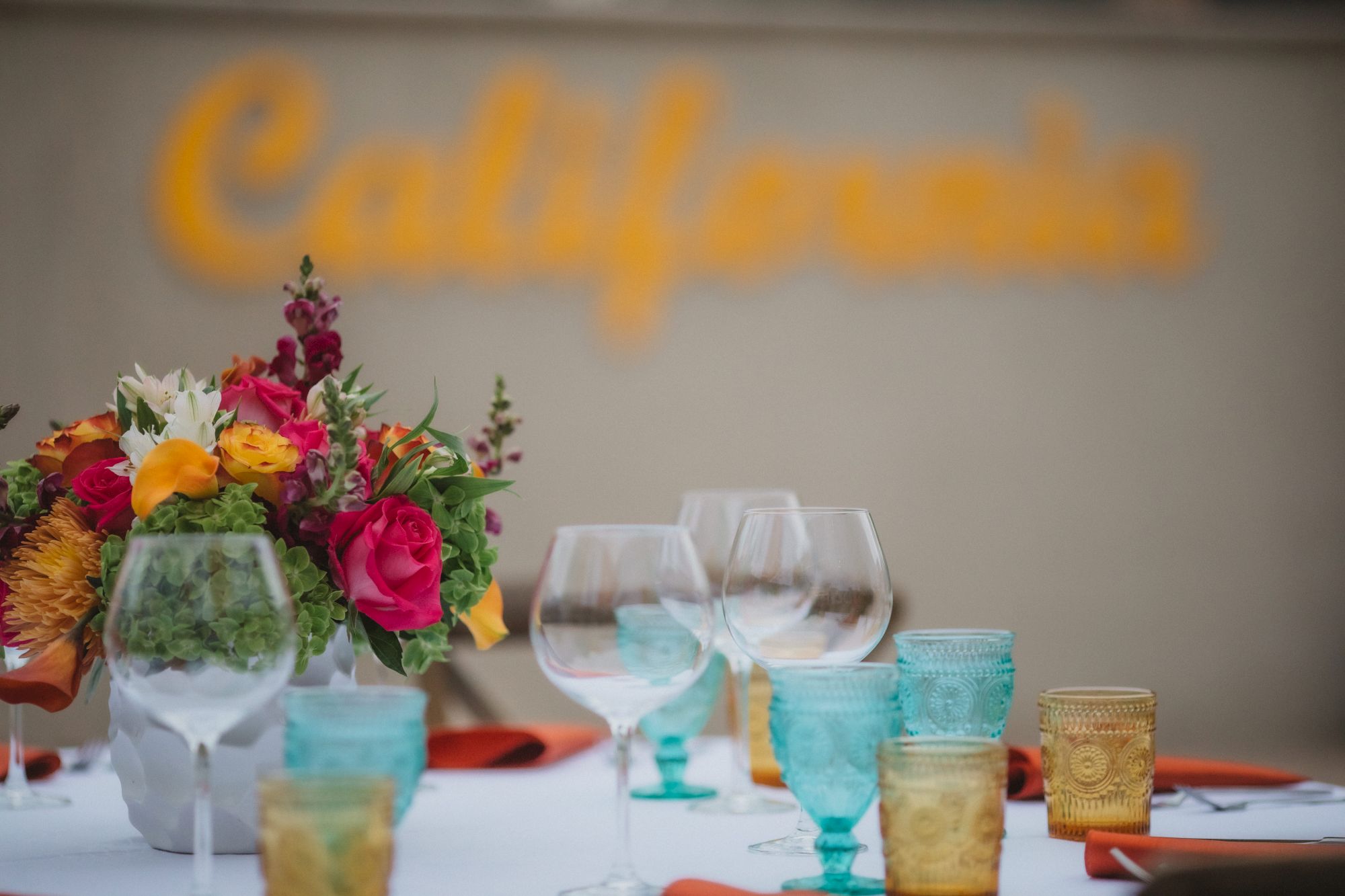 A table setting with colorful glasses, a floral centerpiece, and a blurred "California" sign in the background ends the sentence.