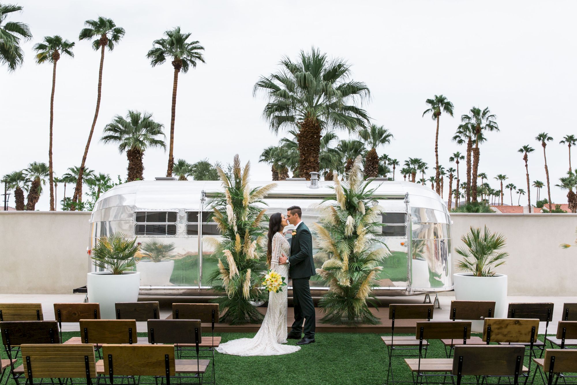 A couple stands in front of an Airstream trailer, surrounded by palm trees and outdoor seating, in a wedding setting.