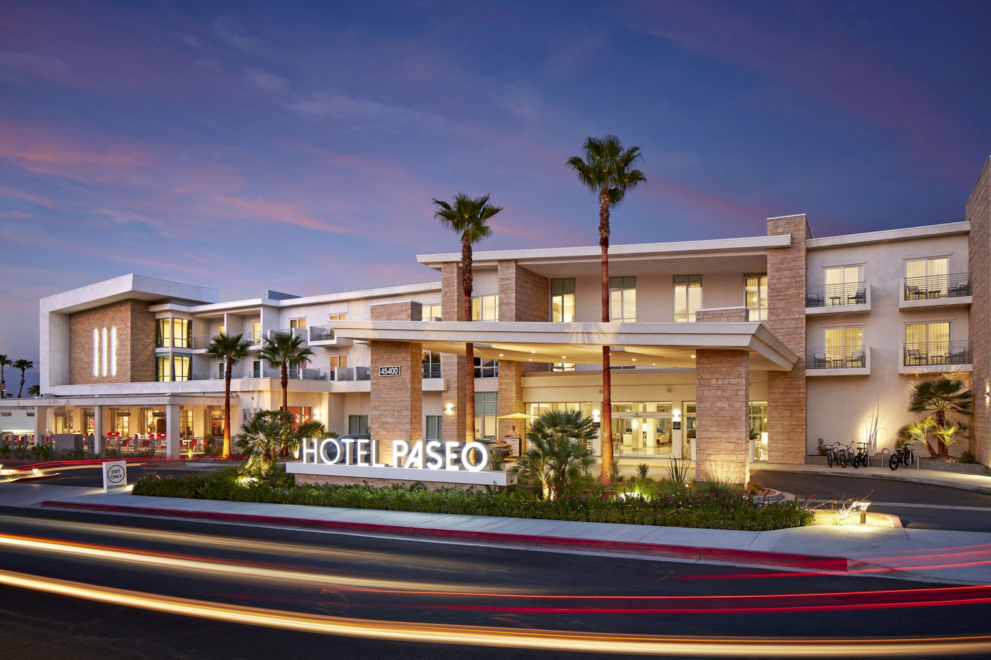 The image shows an elegant hotel exterior with modern architecture, palm trees, and the name “Hotel Paseo” visible at dusk.