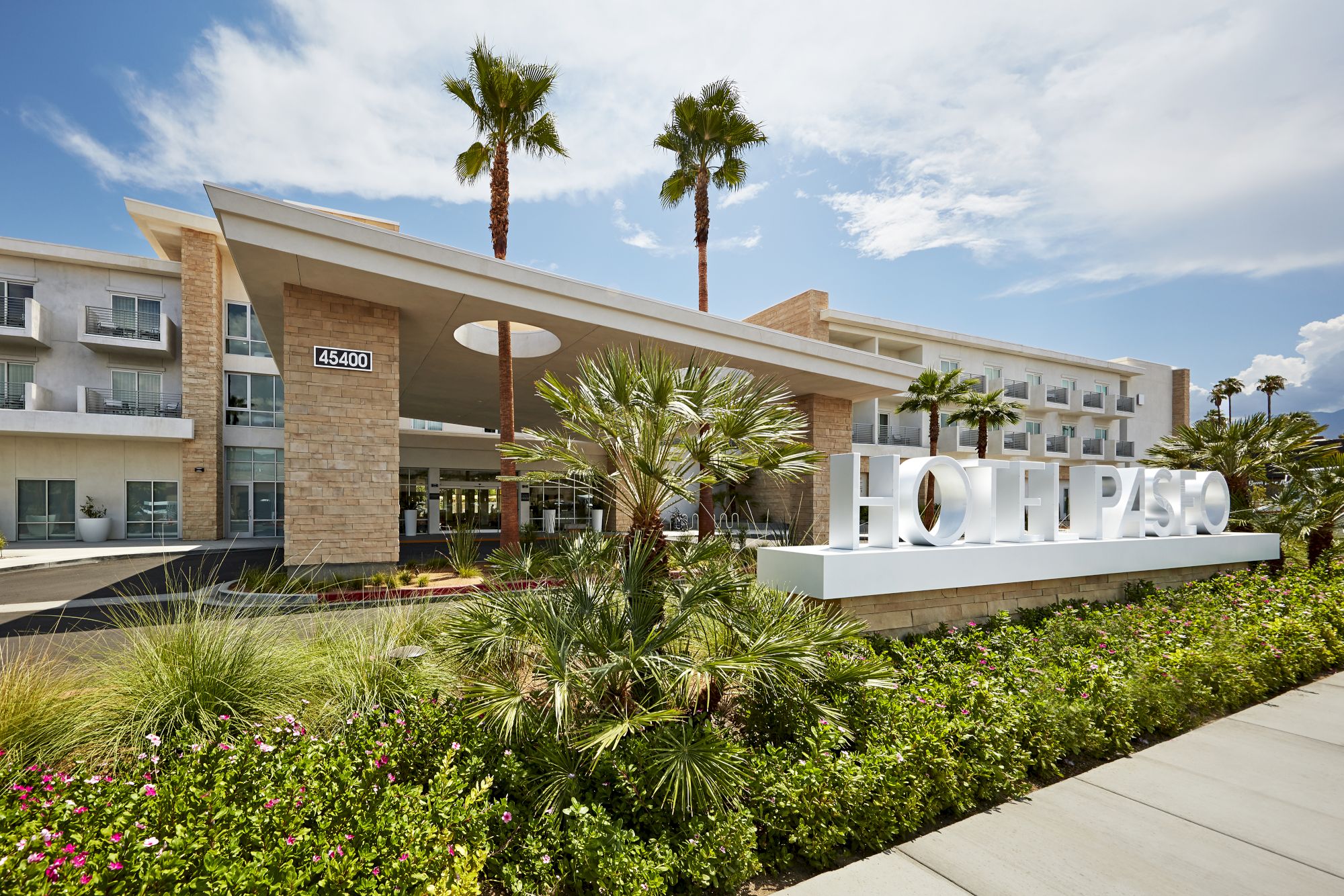 The image shows a modern hotel exterior with palm trees and a clear sky, featuring a prominent entryway and a visible sign.