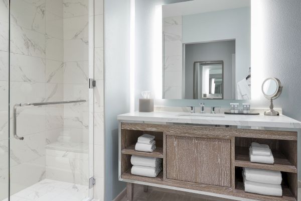 The image shows a modern bathroom with a shower, a large mirror, a sink, and shelves stocked with white towels underneath the vanity counter.