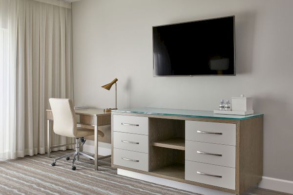 The image shows a modern hotel room with a desk, chair, lamp, TV, and dresser. The decor is minimalistic and neutral in tone.