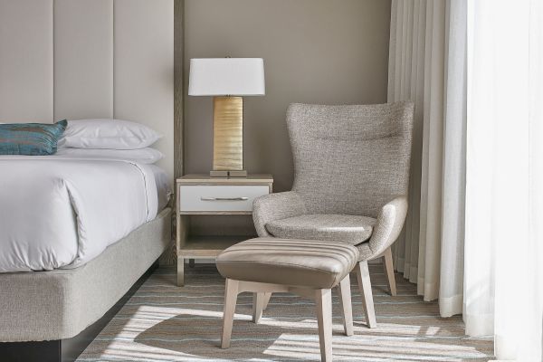 A cozy room with a bed, a nightstand with a lamp, and an armchair with an ottoman, all in neutral tones by a window.