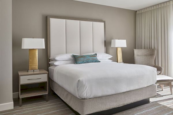 A modern bedroom with a queen bed, two bedside tables with lamps, a chair, and curtains, all in neutral tones, creating a serene atmosphere.