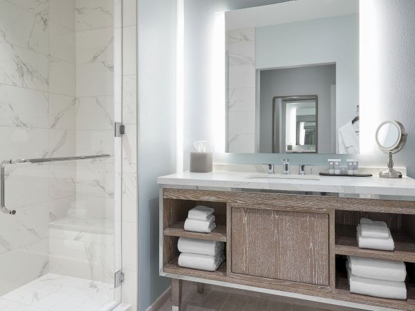 The image shows a modern bathroom with a marble shower, illuminated mirror, and wood vanity stocked with towels and toiletries.