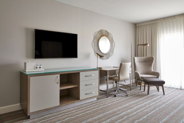 A modern hotel room interior with a TV, desk, chair, wall mirror, and armchair. Bright and minimalistic design with neutral tones.