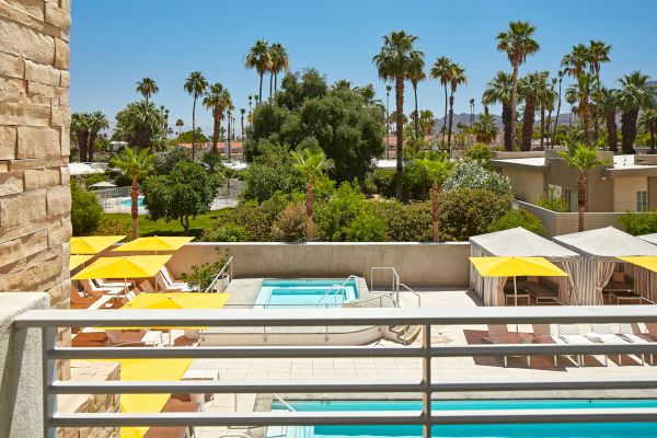 A sunny outdoor pool area with yellow umbrellas, loungers, and cabanas, surrounded by trees and palm trees.