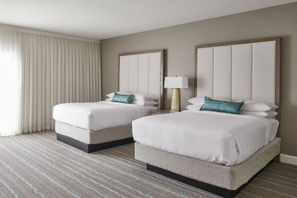 The image shows a hotel room with two double beds, white bedding, teal cushions, a lamp between them, and a large window with curtains.