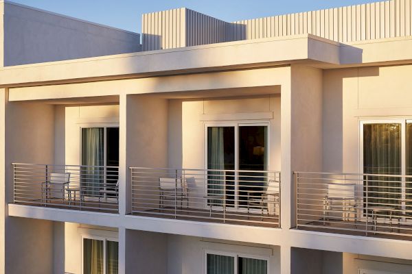 The image shows a modern building with balconies and sliding glass doors, featuring metal railings and outdoor seating.