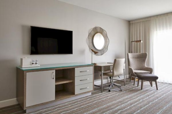 The image shows a modern hotel room with a TV, desk, chair, and stylish mirror on the wall, featuring a minimalist and cozy design.