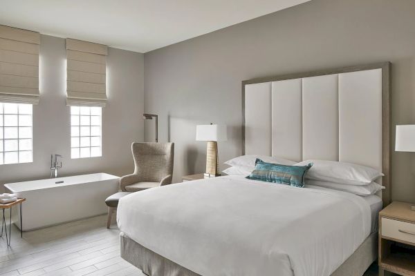 The image shows a modern bedroom with a large bed, a chair, two lamps, a bathtub, and windows, all in a neutral color scheme.