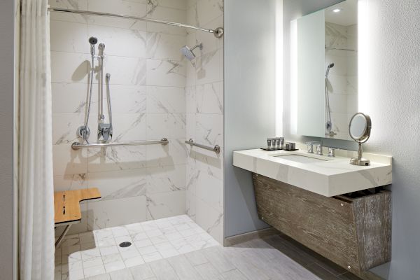 A modern bathroom with a walk-in shower, grab bars, and a wooden bench. Beside it is a vanity with a mirror and a light above.