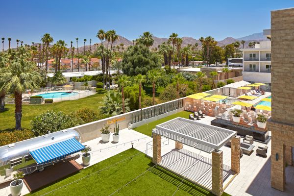 This image shows a sunny outdoor area with palm trees, a pool, lounge areas, and seating, set against a backdrop of mountains and clear skies.