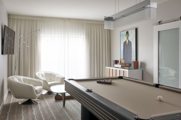 The image shows a modern room with a pool table, white armchairs, a floor lamp, wall art, and a side table against sheer curtains.