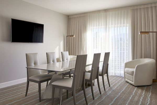 A modern dining room with a glass table, six chairs, a wall-mounted TV, an armchair, and large curtains by a window complete the scene.