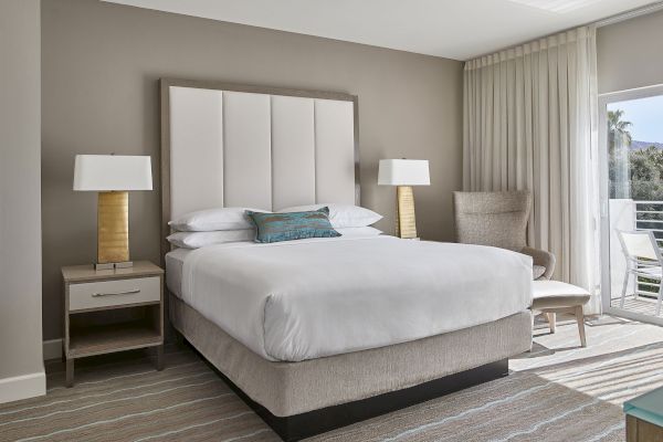 A modern hotel room with a king-sized bed, two bedside tables with lamps, a chair, and curtains partially open to a view outside.