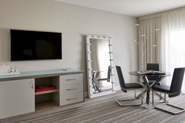 The image shows a modern hotel room with a wall-mounted TV, a large mirror, a dining table with chairs, and sheer curtains.