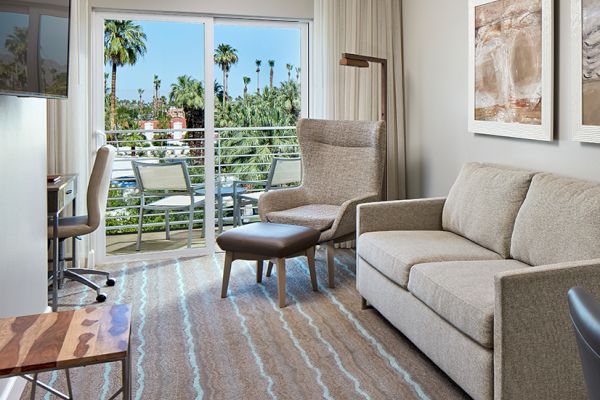 A modern hotel room with a sofa, armchair, desk, and large window showing a balcony with palm trees outside. Art hangs on the walls.