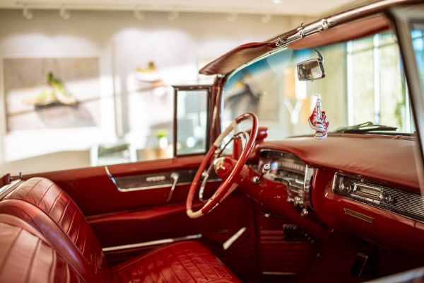 The image shows a vintage car interior with red leather seats and a classic steering wheel. The background displays blurry wall art.