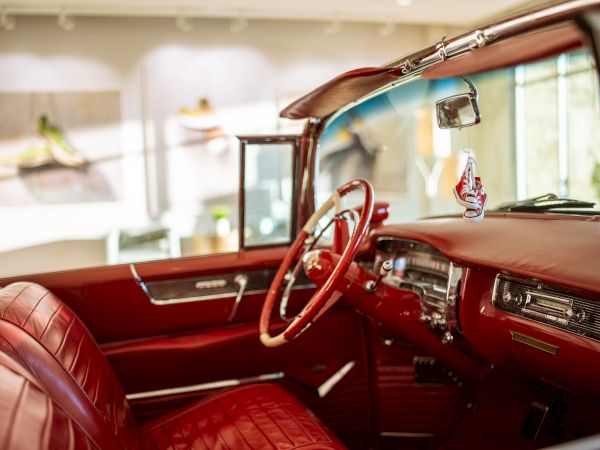 The image shows a vintage car interior with red leather seats and a classic steering wheel. The background displays blurry wall art.