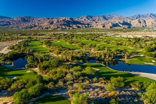 Aerial view of a lush golf course with ponds, surrounded by desert landscape and mountains under a clear blue sky.