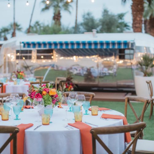 Outdoor dining setup with round tables, colorful decor, and string lights, near a retro trailer and palm trees, creating a festive ambiance.