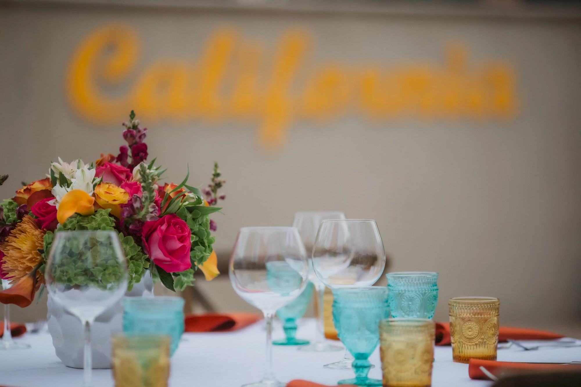 A table set with colorful glassware and a vibrant floral centerpiece, with the word "California" blurred in the background.