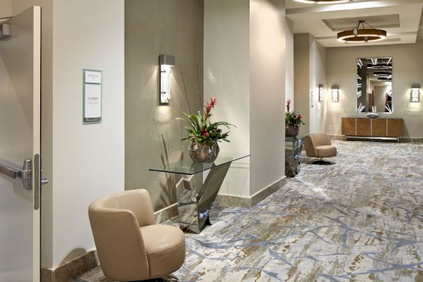 A hotel hallway with stylish decor, featuring a chair, a side table with flowers, wall lights, and patterned carpeting, creating an inviting ambiance.