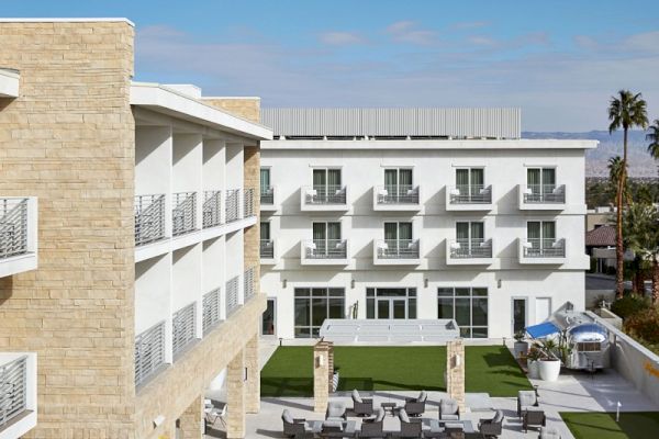 The image shows a modern hotel with multiple stories, featuring a courtyard with green lawns, yellow umbrellas, and outdoor seating areas.