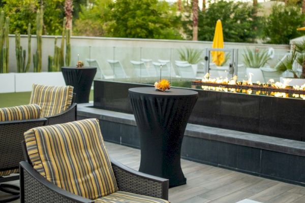 The image shows an outdoor patio with striped cushioned chairs, a black pedestal table, and a fire pit, surrounded by greenery and palm trees.