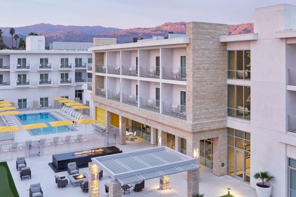 A modern hotel with a pool, outdoor seating area, and a pergola on a green lawn, set against a mountain backdrop under a pastel sky.