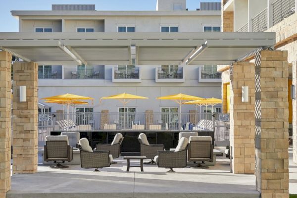 The image shows an outdoor patio with seating, yellow umbrellas, and a modern building in the background, under a clear blue sky.