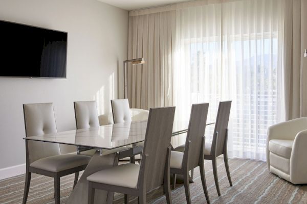 A modern dining room features a glass table, six chairs, a wall-mounted TV, and a large window with sheer curtains.