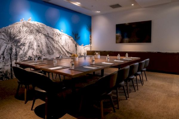 A modern conference room with a long table, chairs, and a large blue mountain mural on the wall. There's also a TV screen.