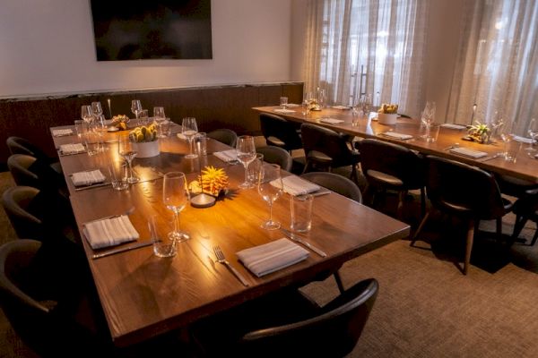 This image shows a dimly lit dining room with a long table set for a meal, featuring wine glasses, napkins, and decorative flowers.