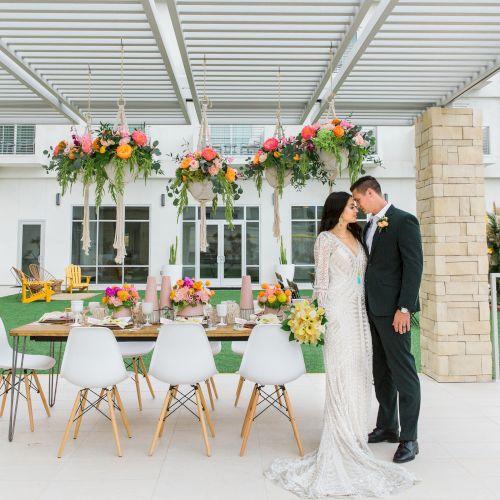 A couple stands in formal attire near a decorated table with floral arrangements. The setting is outdoors with modern architecture.