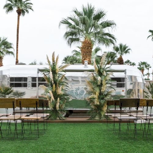 The image shows a vintage trailer with palm trees in the background, surrounded by chairs and greenery.