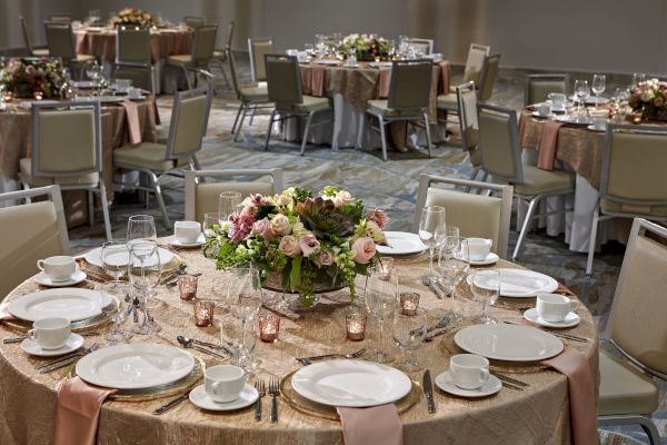 A banquet hall with elegantly set tables, featuring floral centerpieces, plates, glassware, and napkins on beige tablecloths.