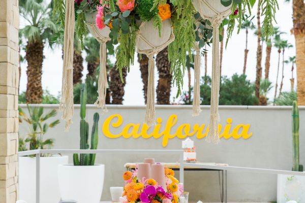 A table set for a meal outdoors, decorated with colorful flowers and greenery. A "California" sign is visible in the background.