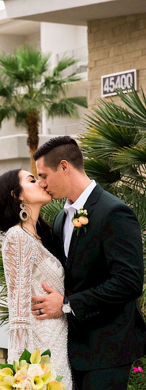 A couple in wedding attire kisses in front of Hotel Paseo, surrounded by palm trees and flowers.