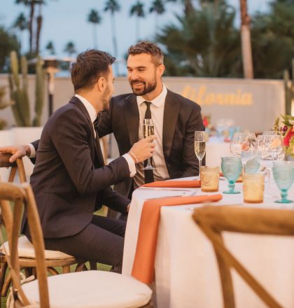 Two people in suits are seated at a decorated outdoor table, engaged in conversation, with flowers and glassware present.