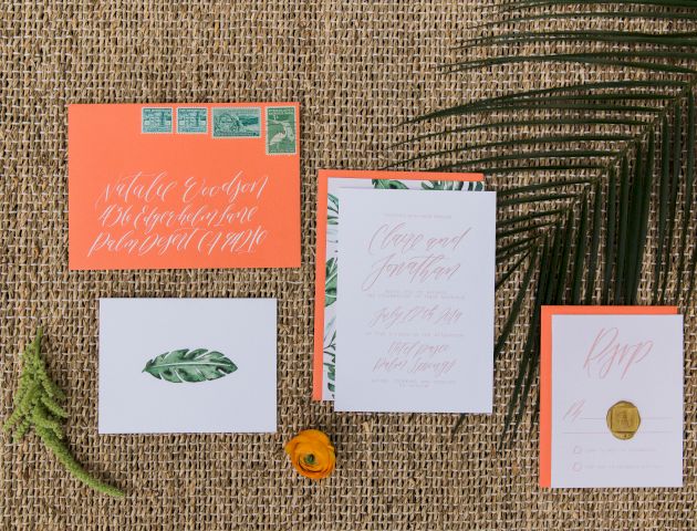 The image shows an invitation set with envelopes, cards featuring leafy designs and calligraphy, placed on a woven mat with greenery nearby.