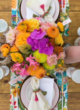 A colorful table setting with floral centerpiece, patterned placemats, plates, napkins, and glasses on a wooden table.