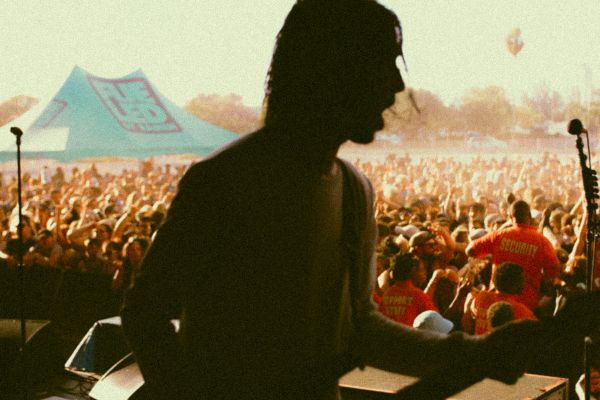 A silhouette of a musician playing guitar on stage with a large audience and festival atmosphere in the background.