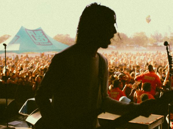 A silhouette of a musician playing guitar on stage with a large audience and festival atmosphere in the background.