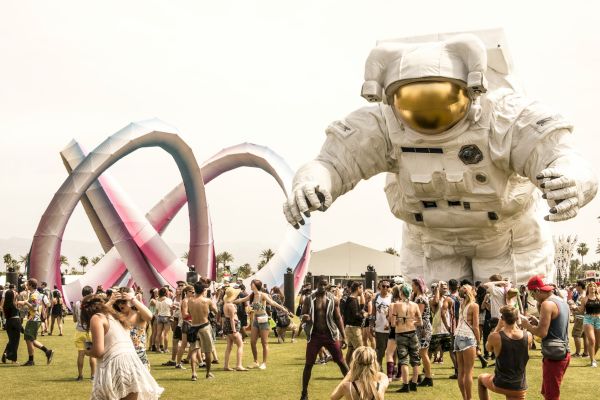 A festival scene with a large astronaut sculpture, colorful structure, and people enjoying the outdoor event under a sunny sky.