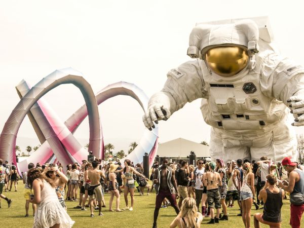 A festival scene with a large astronaut sculpture, colorful structure, and people enjoying the outdoor event under a sunny sky.