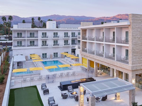 A modern hotel with a pool, yellow umbrellas, seating areas, and a small golf putting green, set against a mountainous backdrop.